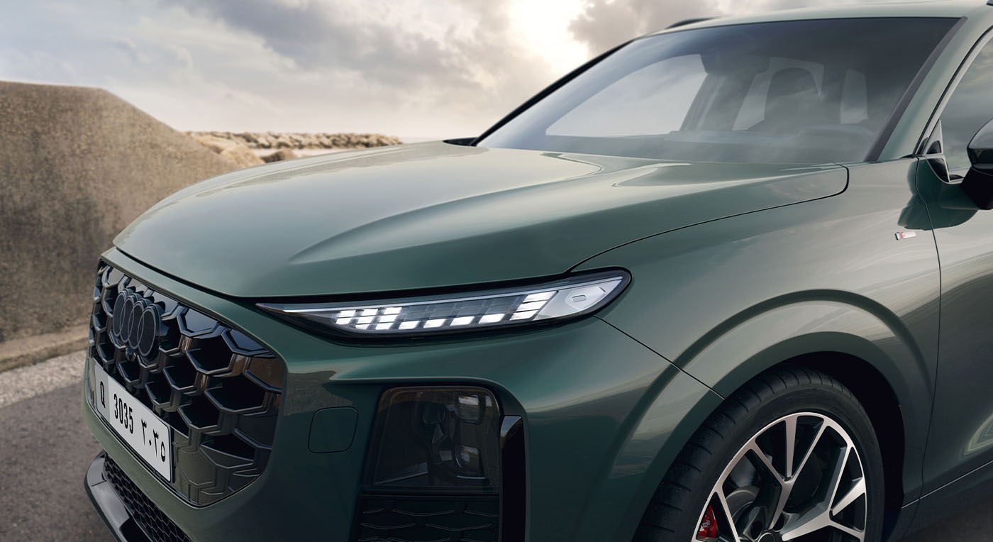 A sleek green Audi Q3 SUV parked by the seaside under a cloudy sky. The focus is on the front headlight and grille, highlighting the vehicle's modern design.